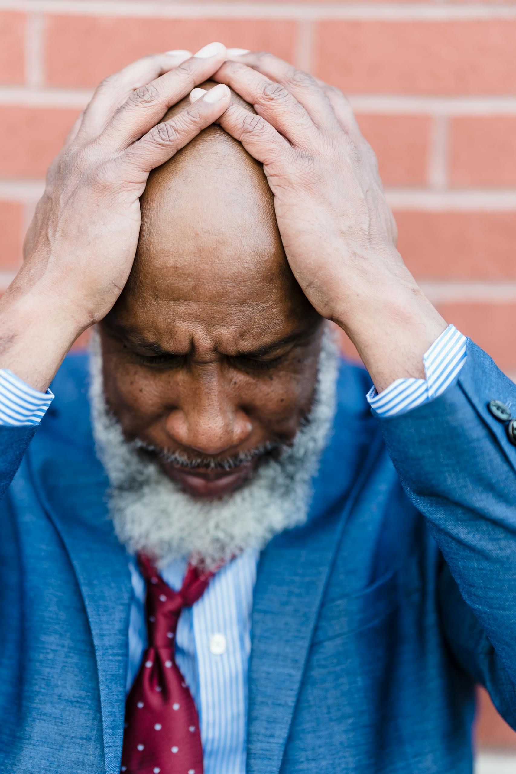Worried bearded African American male entrepreneur in formal clothes standing near brick wall with hands on head after hard workday